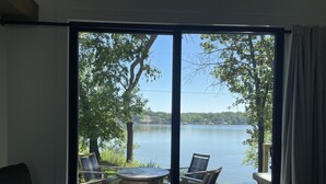Una habitación con un gran ventanal corredero que da a un espacio exterior con mesa y sillas, con vista a un lago, árboles y edificios a lo lejos.