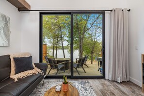 A modern living room with a sliding glass door leading to a deck with a table and chairs, a black leather sofa, and a small coffee table with a plant. - Lakeside Modern Luxury w/Lake Views (Grove)