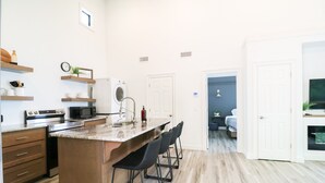 Una cocina moderna con una isla central, taburetes de bar, electrodomésticos integrados y vistas al dormitorio contiguo.