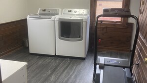 Une buanderie équipée de deux machines à laver et de deux sèche-linge à chargement frontal, d’un tapis de course et d’une porte en bois donnant sur l’extérieur.