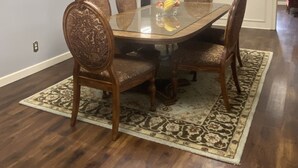 Une salle à manger avec une table à plateau de verre, entourée de chaises décorées, un tapis à motifs et un tableau encadré accroché au mur.