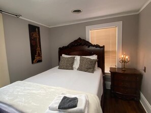 A neatly made bed with a white comforter and two patterned pillows, a wooden headboard, a wall-mounted Eiffel Tower artwork, a bedside table with a candelabra, and a window with blinds. - YOUR AMAZING 4 BEDRROMS GREAT PRICE (Willmar)