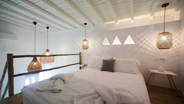 Una camera da letto con una testiera in legno, un letto grande con lenzuola bianche, due lampade pendenti in vimini e un comodino con una lampada bianca.
