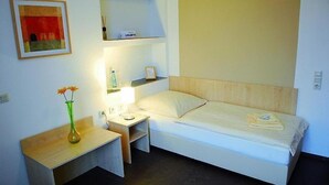 A small, neatly arranged bedroom with a single bed, a bedside table with a lamp, a small shelf above the bed, and a framed picture on the wall. - Adam Hotel - Giesen (Giesen)