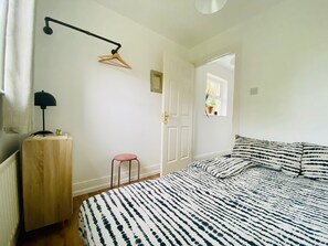 A bedroom with a bed, a wooden nightstand, a hanging light, a door, and a window. - The Yellow Door Cottage (Moville)