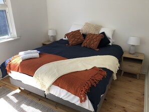 A neatly made bed with a blue and white duvet, brown and white throw blankets, and several pillows. There are two bedside tables with lamps and a small shelf. - Absolute Beachfront - simply enjoy  (Henley Beach)