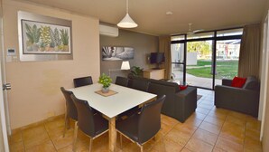 Un coin repas avec une table blanche, des chaises noires et une plante en pot. On aperçoit un canapé et un téléviseur à l’arrière-plan.
