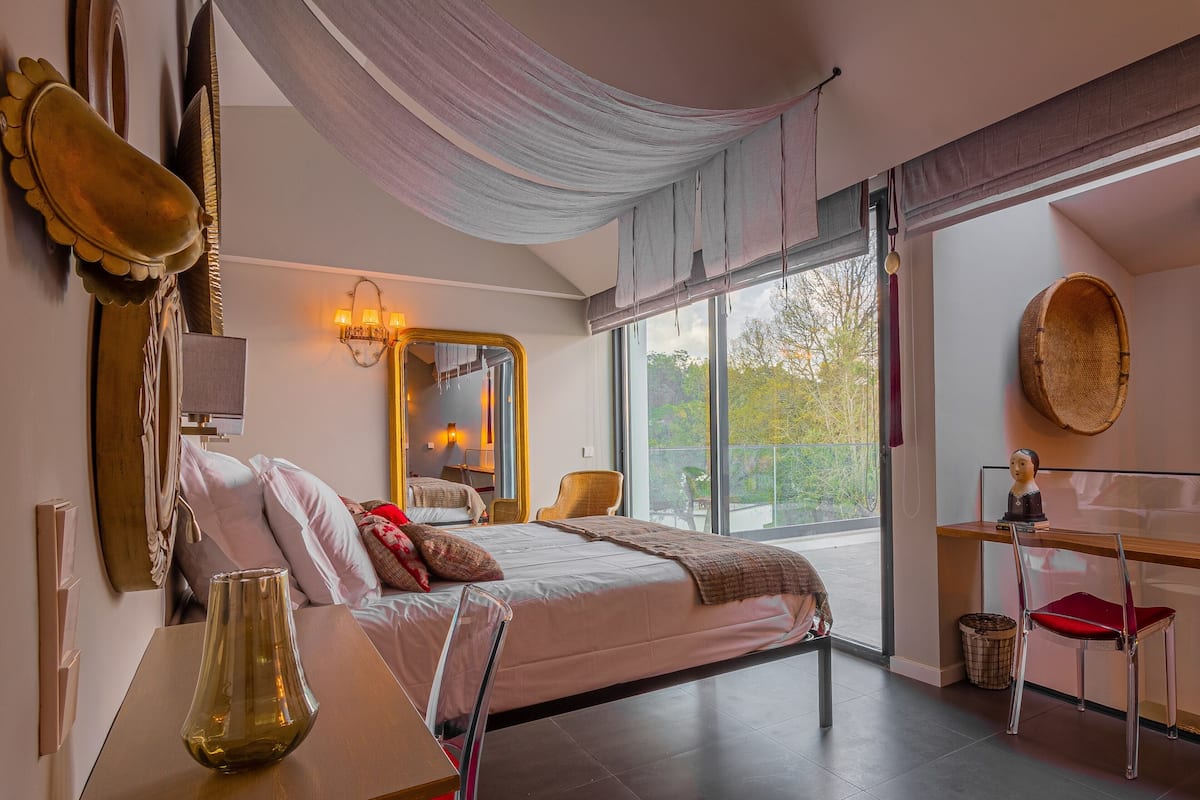 A modern bedroom with a large bed, a desk, and a chair. There is a window with a view of trees and a balcony.