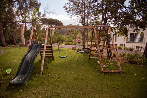 Un parque infantil con tobogán, columpios y estructura para escalar en un área de césped con árboles y un edificio al fondo.