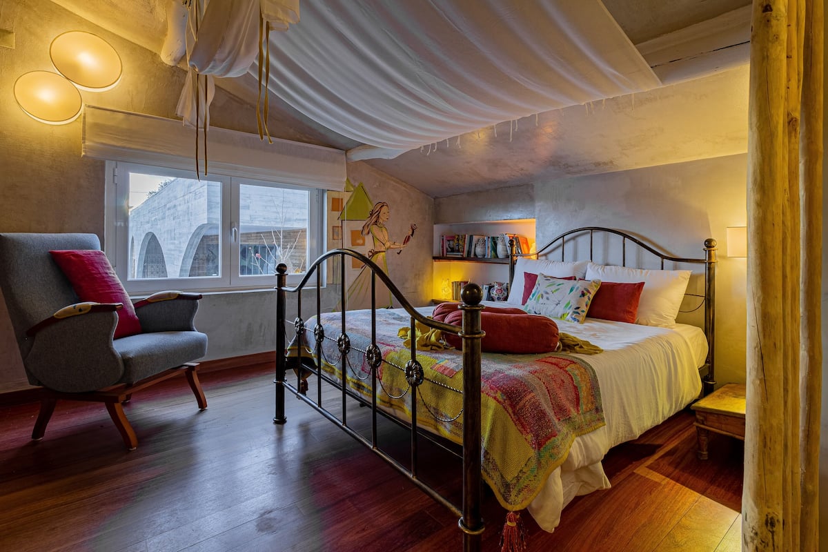 A bedroom with a bed, a chair, a window, and a bookshelf.
