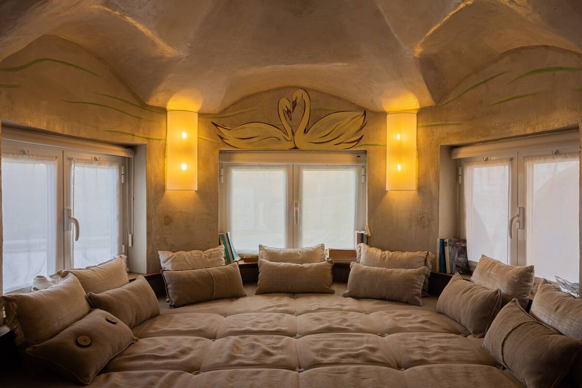 A large, centrally positioned bed with multiple pillows, flanked by windows with curtains, and wall-mounted lighting fixtures.