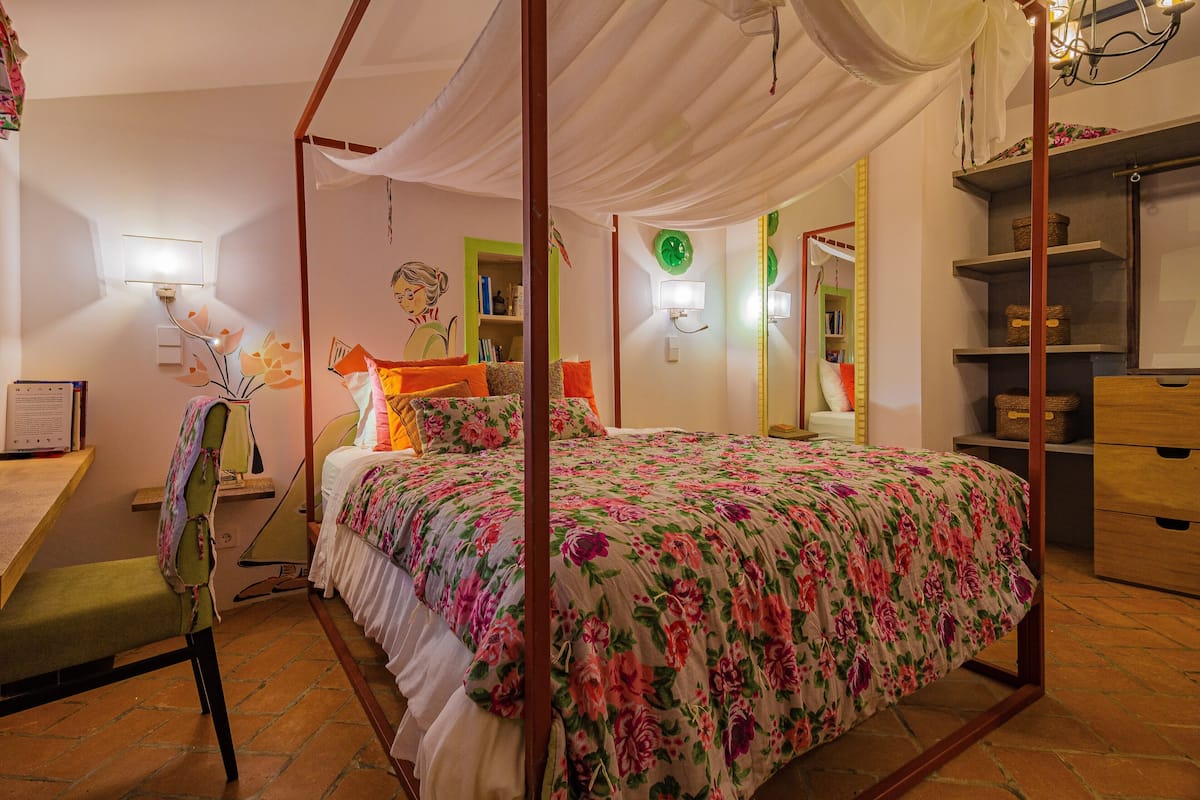 A bedroom with a four-poster bed, a desk with a chair, a bookshelf, and a mirror.