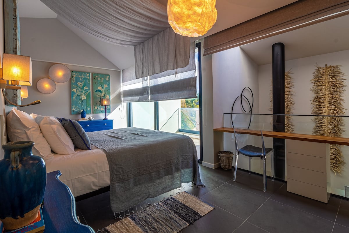 A bedroom with a bed, bedside table, lamp, and a view of the outdoors.