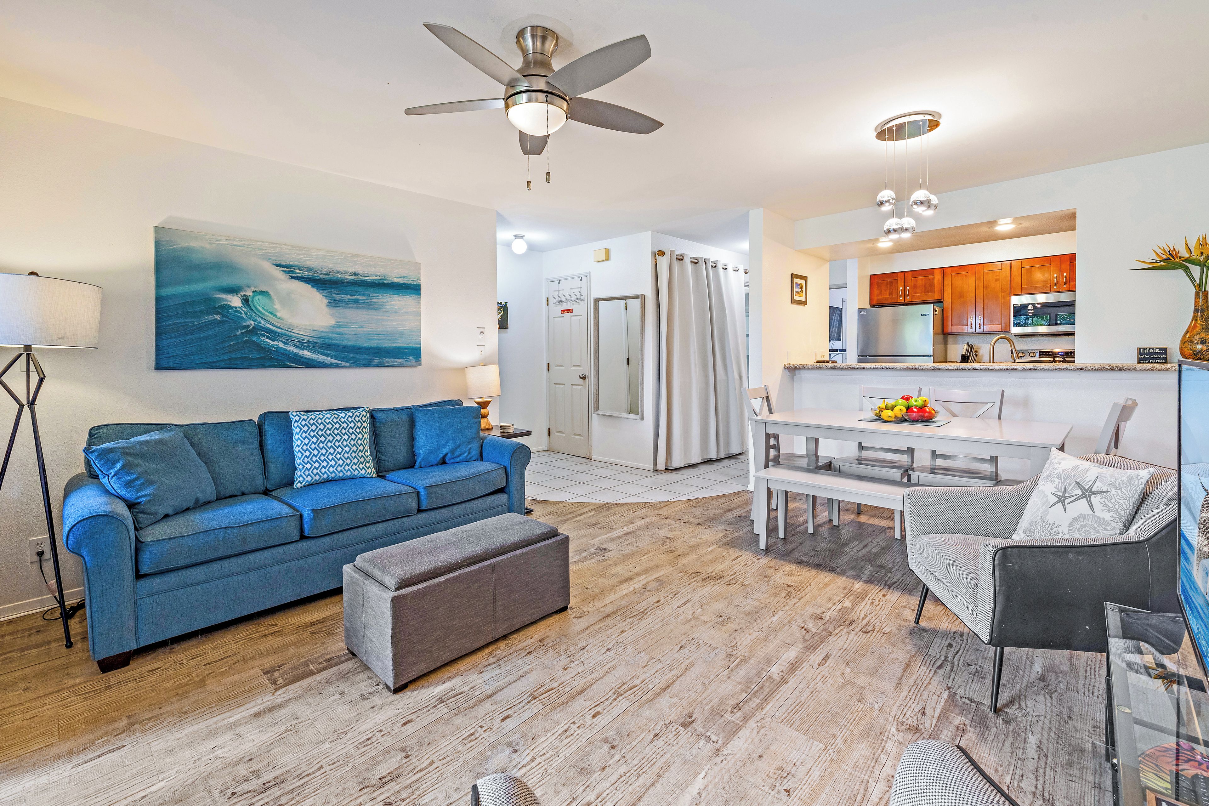 A modern living room with a blue sofa, a grey ottoman, and a dining area with a white table and chairs. A kitchen is visible in the background with wooden cabinets and stainless steel appliances.