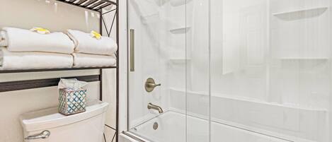 A bathroom with a glass-enclosed shower, a toilet, and a shelf with folded towels.