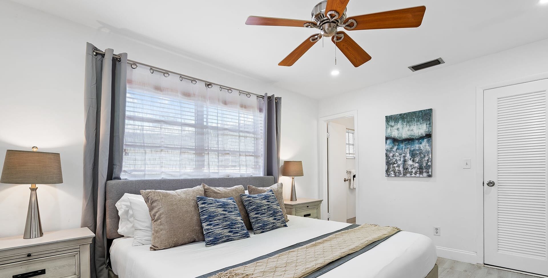 A bedroom with a bed, two bedside tables, a ceiling fan, a window with curtains, a painting, and a door.