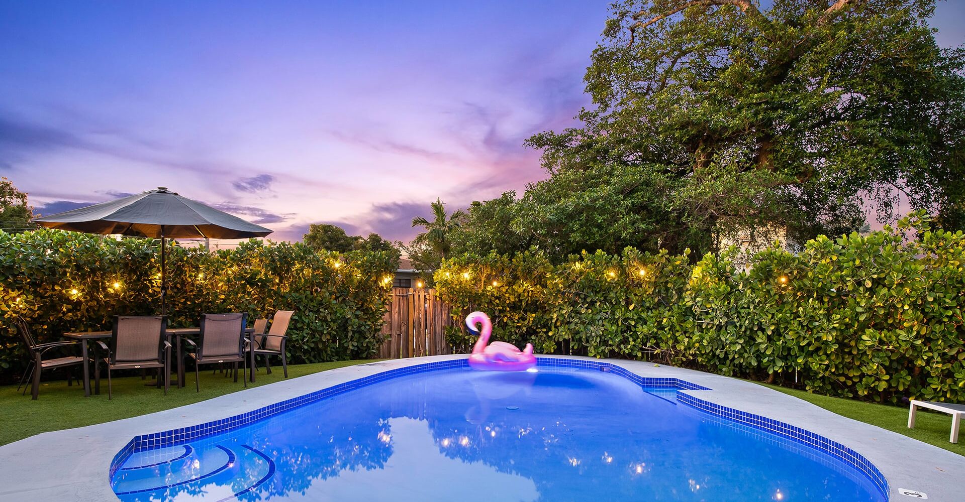 A swimming pool with a pink flamingo float, surrounded by a wooden fence, chairs, and string lights.