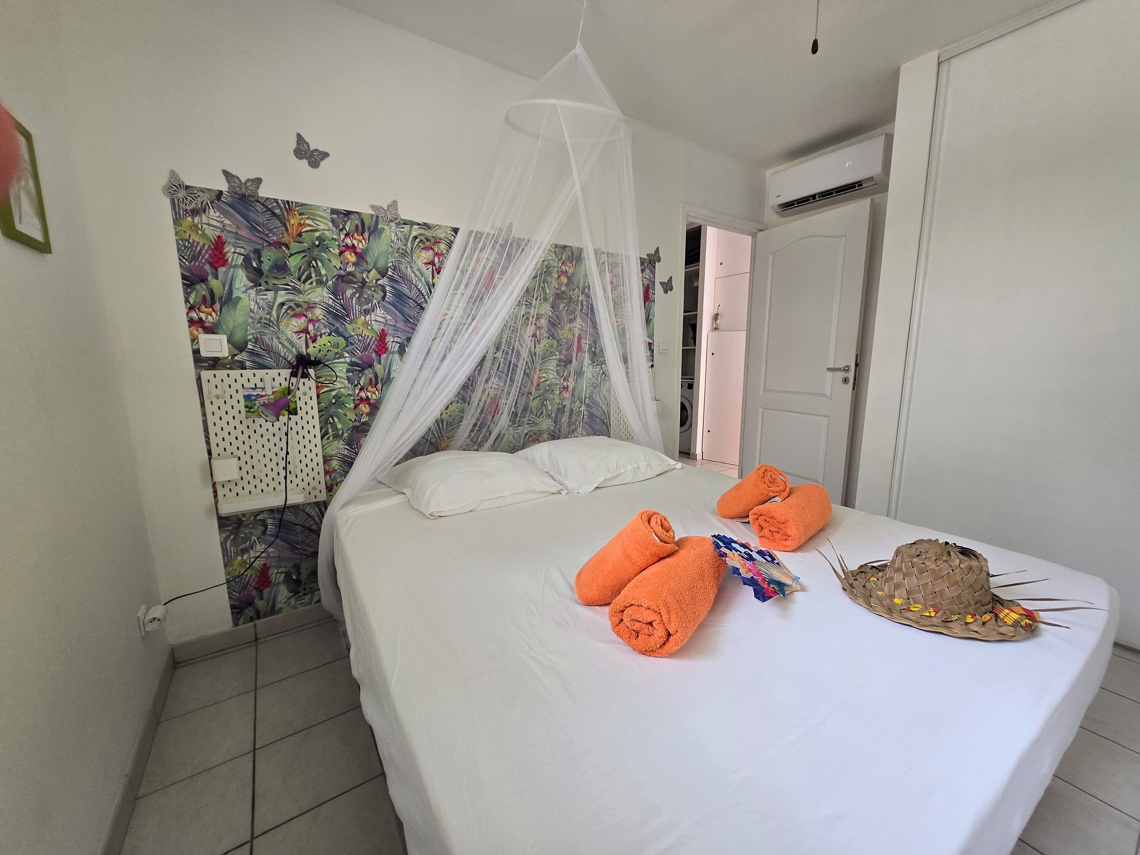 A neatly made bed with white linens and orange towels, a mosquito net above, and a colorful tropical-themed wall art.