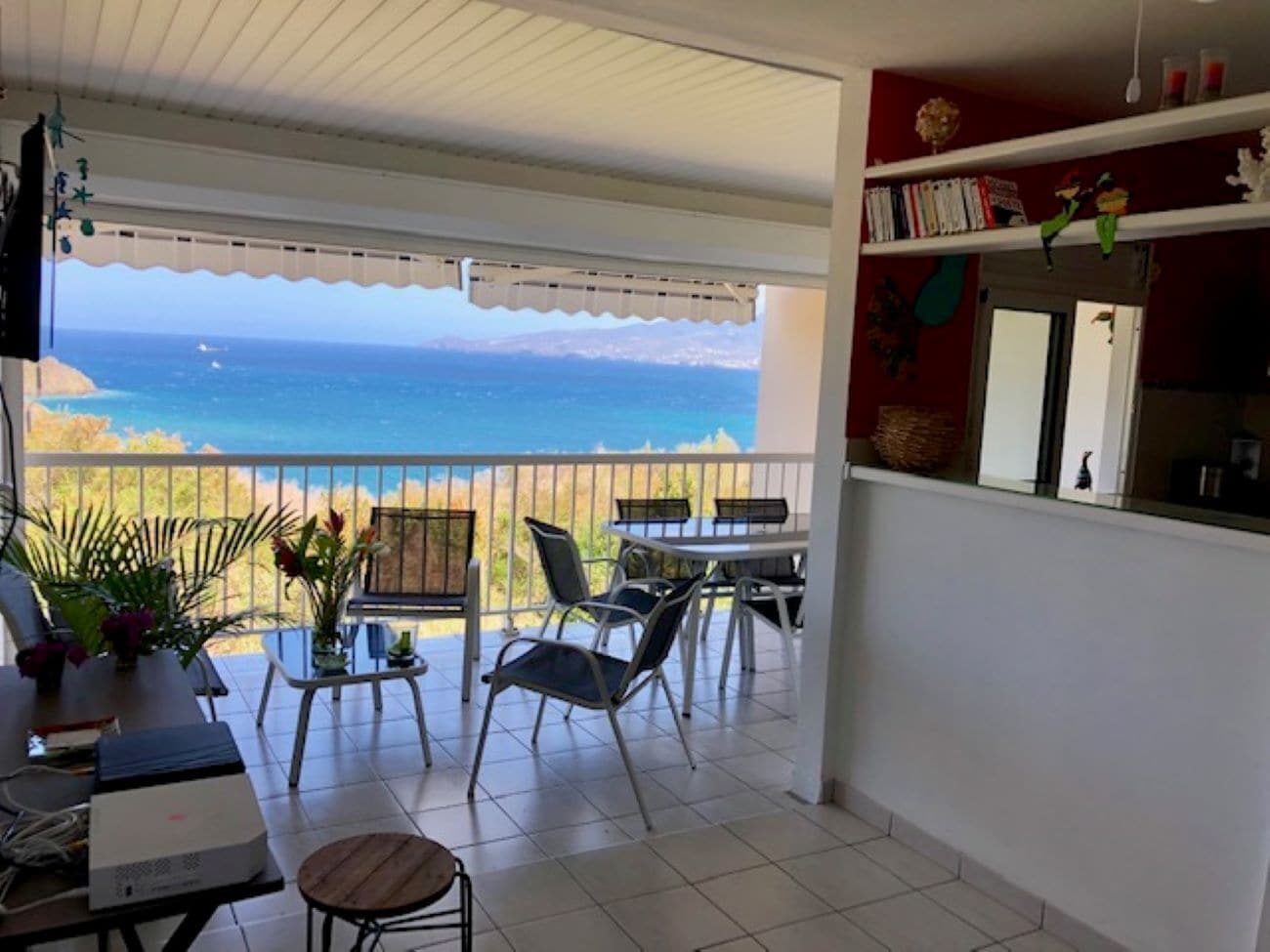 En balkon med udsigt over havet, et spiseområde med stole og et bord, samt en reol med bøger og pynteting.