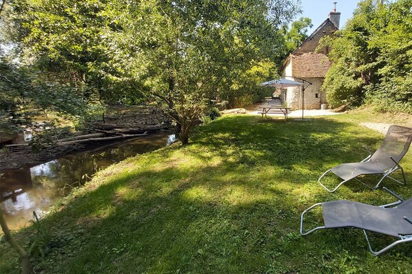 Un espace extérieur paisible, avec une pelouse verdoyante, un petit étang et un pont de pierre. On aperçoit deux chaises longues et un kiosque au fond.