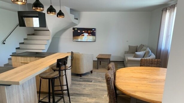 Una cocina moderna con una isla de madera, taburetes de bar, una mesa de comedor con sillas, una escalera y un televisor montado en la pared.