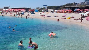 En strand med klart vand, parasoller og mennesker der svømmer og solbader.