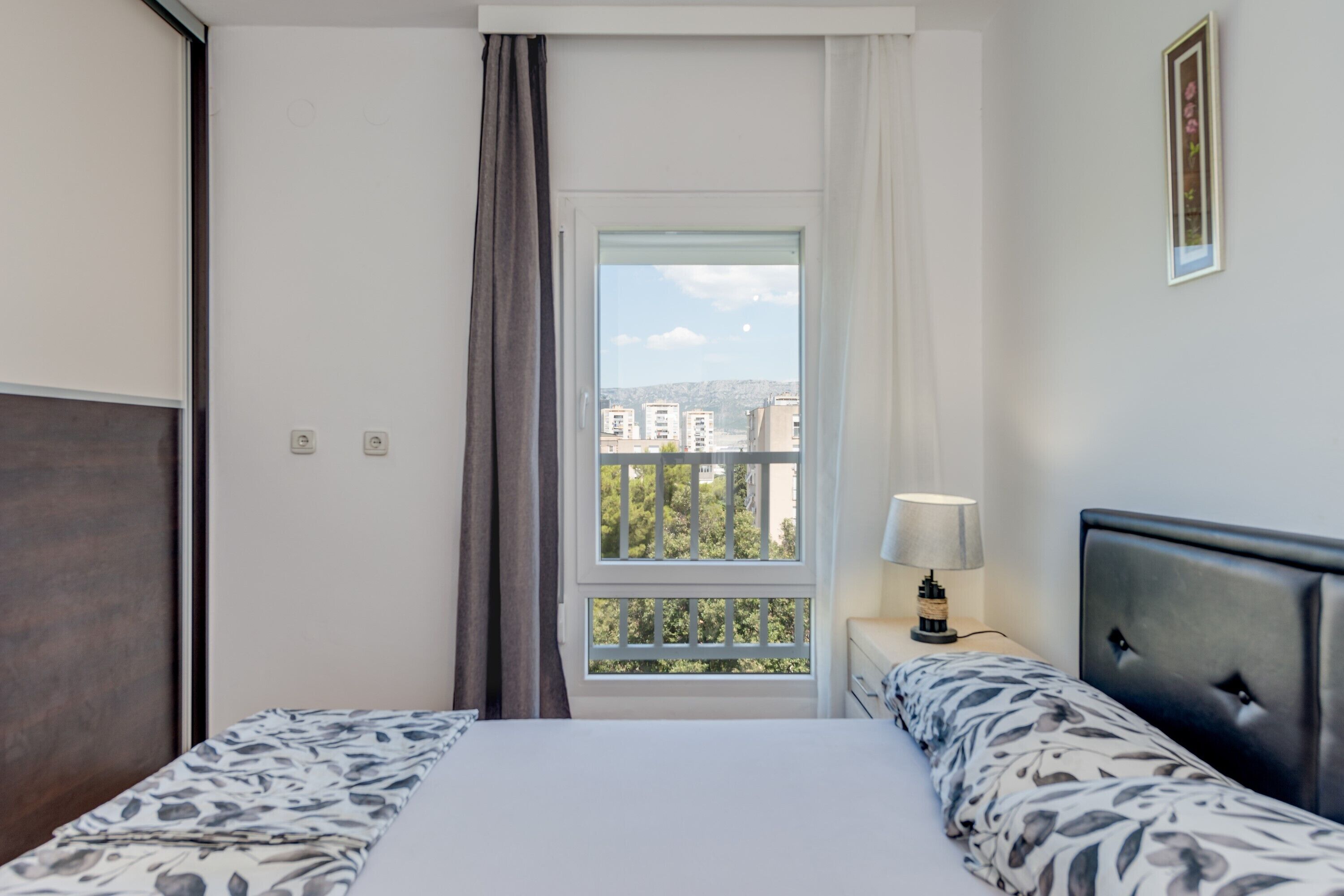 Habitació d'hotel amb un llit, una nitòria, una làmpada, una finestra amb vistes a edificis i una cortina.
