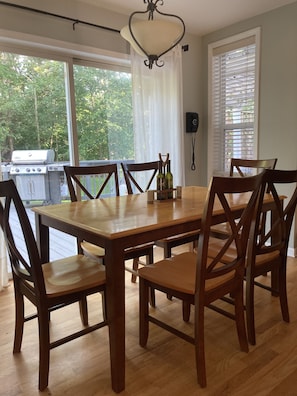A dining area with a wooden table and chairs, large windows with curtains, and a view of a grill outside.