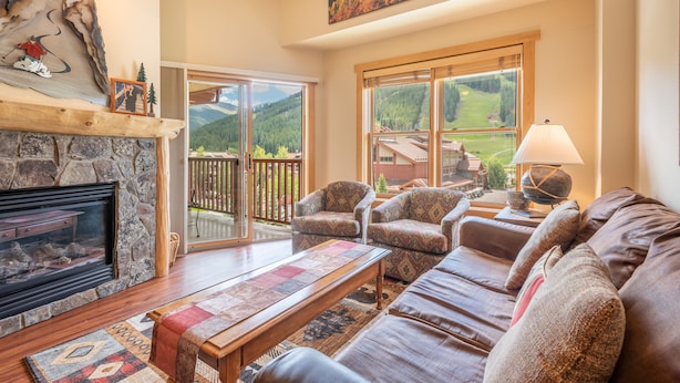 A cozy living room with a stone fireplace, comfortable seating, a large window with a scenic view, and a wooden ceiling. - Perfect views of the Super Bee Lift, Free WiFi, underground parking (Copper Mountain)