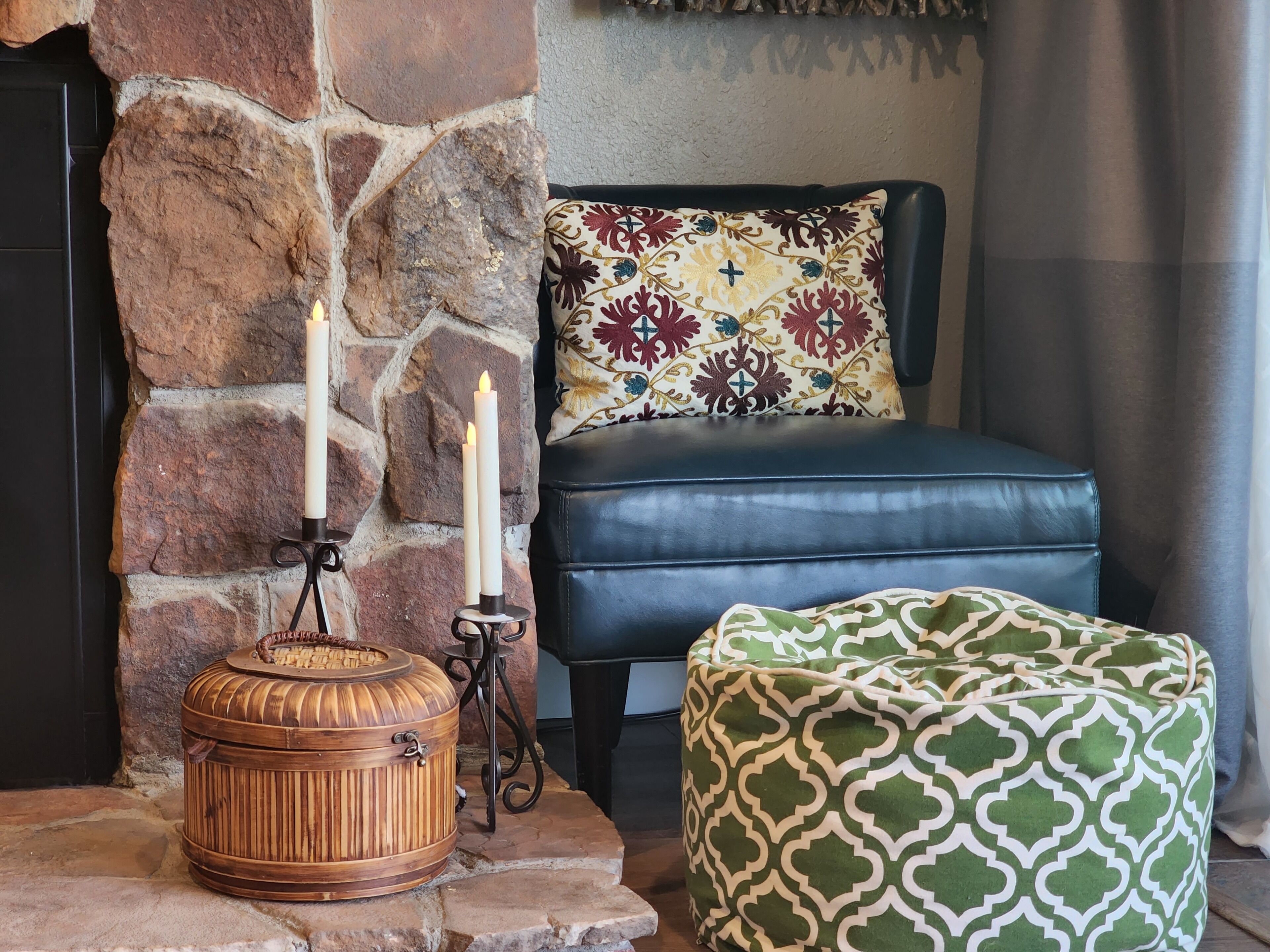 Une cheminée en pierre avec trois bougies, une malle en bois, un canapé en cuir noir, un coussin à motifs et un pouf vert.