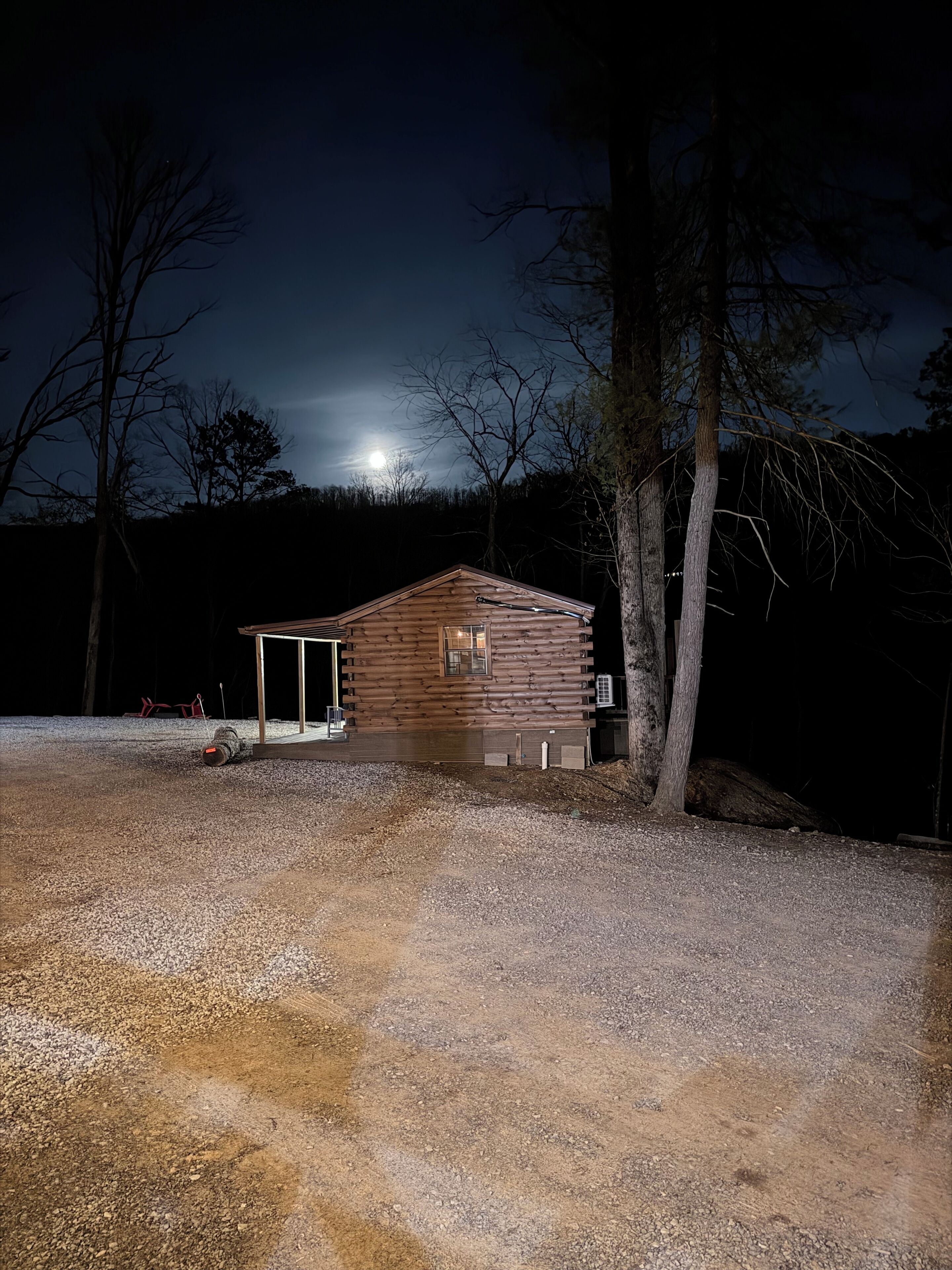 Eine Holzblockhütte mit Veranda und einem Schotterweg, umgeben von Bäumen und einem dunklen Himmel.