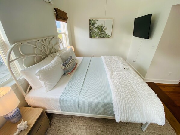 Una cama bien hecha con cabecera blanca, colcha azul claro y manta blanca. Se aprecia un pequeño tocador de madera con una lámpara y una concha decorativa. Encima de la cama, hay un cuadro enmarcado en la pared.