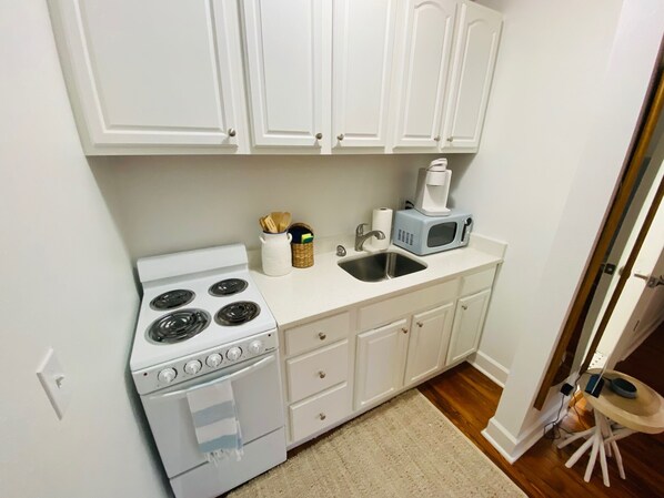 A compact kitchen with white cabinets, a stove, an oven, a microwave, a sink, and a small table with a chair.