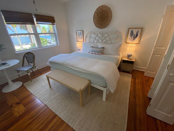 A bedroom with a large bed, a bedside table, a chair, a window with blinds, and a view of palm trees.