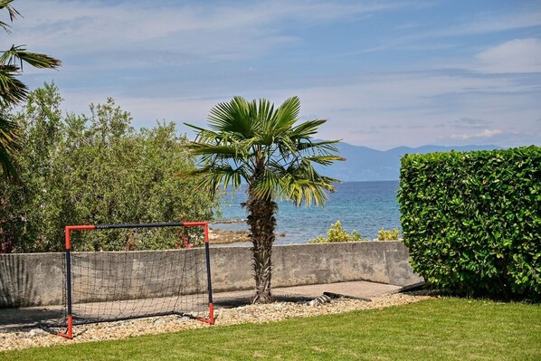 A netted soccer goal in a grassy area with a palm tree and a hedge in the foreground, and a body of water with mountains in the background.