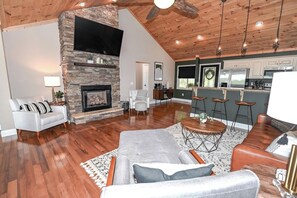 A spacious living room with a fireplace, a television mounted on a stone wall, and a kitchen area with bar stools.