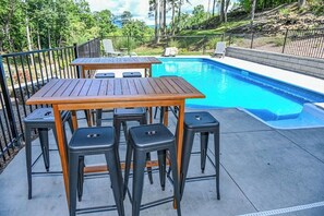 Zona de piscina con mesa de madera y taburetes negros, rodeada por una valla metálica.