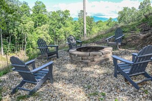 A circular seating arrangement around a stone fire pit with Adirondack chairs, set on a gravel surface with a forest backdrop. - Barefoot in the Blue Ridge (Hayesville)