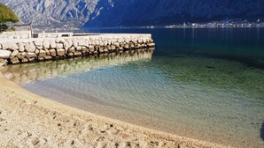 Eine Küstenansicht mit freiem Blick auf einen felsigen Wellenbrecher, der zu ruhigem Wasser, einem Sandstrand und einer gebirgigen Kulisse führt.
