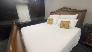 A neatly made bed with white linens and two decorative pillows, a wooden headboard, and a dark wooden dresser with a mirror.