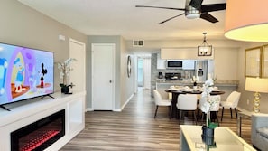 A modern living room with a flat-screen TV, a fireplace, a dining area with a table and chairs, a kitchen in the background, and a ceiling fan. - Grand Deluxe Garden 3BR Penthouse near Disney Parks (Kissimmee)