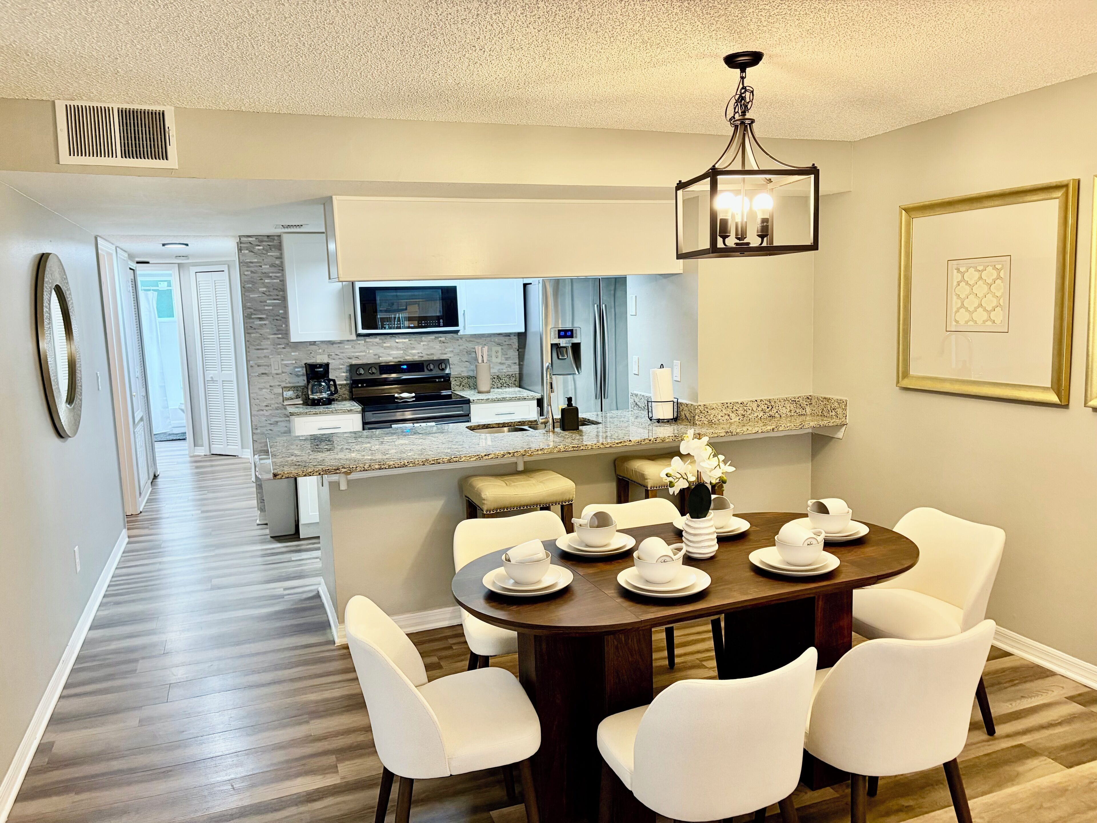 A modern kitchen with a microwave, stove, and coffee maker, a dining area with a table set for four, and a hallway leading to a bedroom.