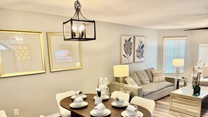 A modern living room with a dining area, a sofa, a coffee table, and framed artwork on the walls. - Grand Deluxe Garden 3BR Penthouse near Disney Parks (Kissimmee)