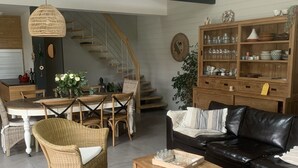 A modern living room with a dining area, featuring a staircase, a black leather sofa, wicker chairs, and a wooden coffee table. - Agréable Maison Bois (Andernos-les-Bains)
