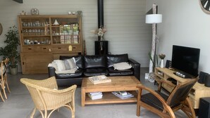 A cozy living room with a black leather sofa, a wooden coffee table, a wicker chair, and a TV stand with a flat-screen TV. - Agréable Maison Bois (Andernos-les-Bains)