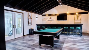 Ein Billardtisch in einem Raum mit hoher Decke und freiliegenden Balken, ein Barbereich mit Hockern und ein großes Fenster mit Vorhängen.