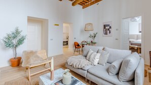 Amplia sala de estar con techo de madera alto, un sofá gris, una pequeña silla de madera, una planta en maceta y un comedor al fondo.