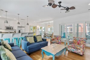 A spacious living room with a blue sofa, a coffee table, and a colorful armchair. A kitchen with white cabinets and a microwave is visible in the background. - Ocean View / Sleeps 14 / Sunshine By The Sea / Grand Pavilion Pool Access (Isle of Palms)