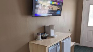 A hotel room with a television mounted on the wall, a small wooden desk with a coffee maker, a microwave, and a trash bin. - The Greenland Motel - Room No. 7 (Munising)