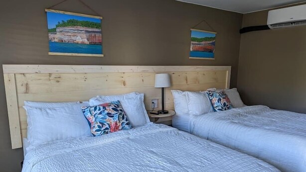 A hotel room with two beds, wooden headboard, white bedding, a nightstand with a lamp, and two wall-mounted paintings. - The Greenland Motel - Room No. 7 (Munising)